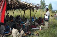 A community are gathered under a shelter listening to a health professional talk on health messages.